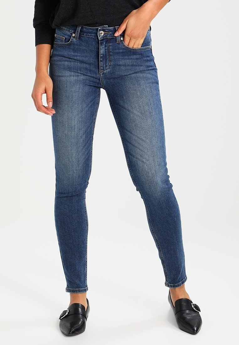 Jeans ajustados azules de mezclilla, con una cintura de altura media, diseño clásico de cinco bolsillos y costuras sutiles. Combinados con bailarinas negras.