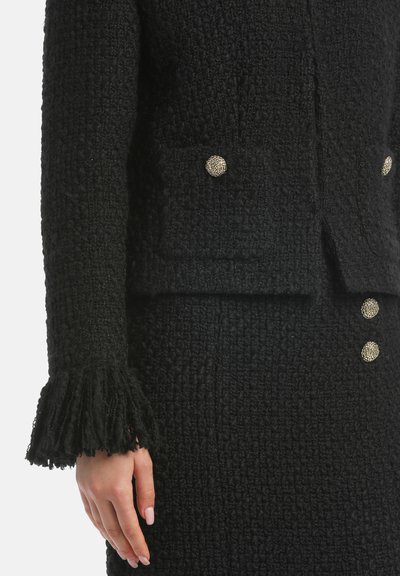 Abito nero con texture, composto da una giacca con due tasche frontali e dettagli in metallo sui bottoni; gonna con bottoni coordinati e frange decorative.
