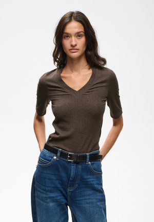 Femme aux cheveux bruns ondulés portant un haut marron à manches courtes en V, rentré dans un jean bleu avec une ceinture noire, debout les mains dans le dos.