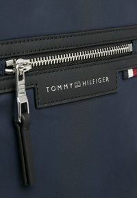 Zaino in nylon blu scuro con una tasca zip nera, accessori in metallo argentato e un'etichetta nera con testo bianco e accenti colorati.