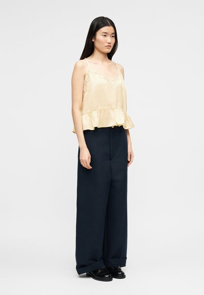 Femme portant un caraco en satin jaune clair avec bordure en dentelle et ourlet à volants, associé à un pantalon large bleu marine foncé et des chaussures noires.