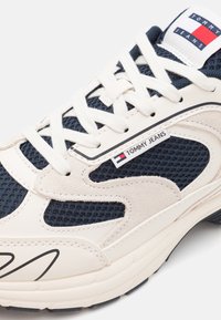 Zapatillas atléticas en blanco y marino que cuentan con materiales de malla y cuero, superficie texturizada, acentos contrastantes y etiqueta de marca.