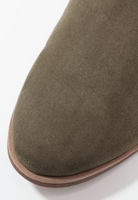 Gros plan sur l'embout d'une chaussure en daim vert olive avec une semelle empilée marron clair sur fond blanc.