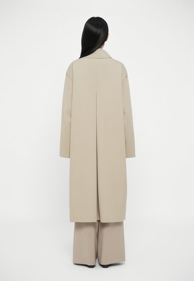 Beige παλτό με γιακά και μακριά μανίκια, με ίσια γραμμή και λεπτομέρεια ραφής στην πλάτη, συνδυασμένο με αντίστοιχους φαρδύς παντελόνες.