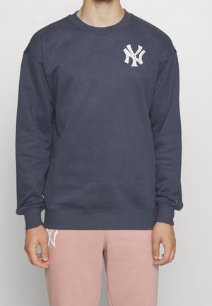 Persona che indossa una felpa blu navy con logo bianco sul petto e pantaloni rosa chiaro con logo abbinato sulla coscia su sfondo uniforme.