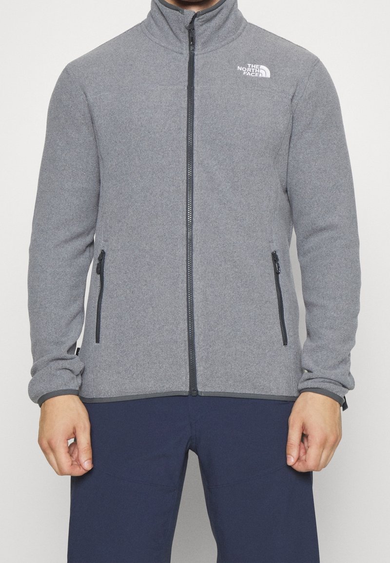 Gray fleece jacket with a high neck, front zipper, two side pockets, and brand logo on the chest. Smooth texture and fitted design.