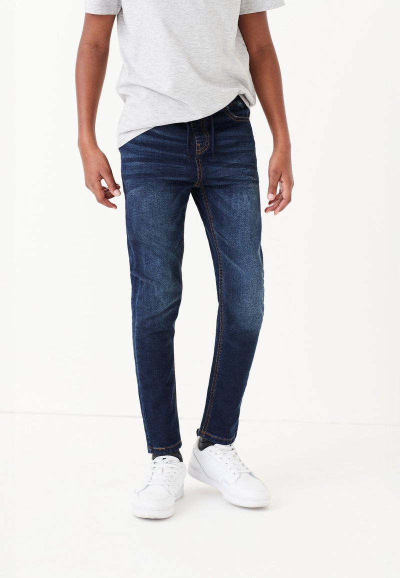 Personne portant un jean slim bleu foncé, un t-shirt gris clair et des baskets blanches, debout devant un fond blanc uni.