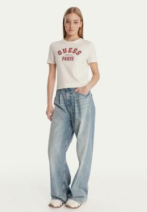 Giovane donna in piedi che indossa una maglietta bianca "Guess Paris", jeans larghi azzurri chiari e sneakers bianche, con la mano in tasca.