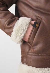 Giacca di pelle marrone con polsini e foderi in fleece bianco, dotata di tasche laterali e un'etichetta nera con testo giallo. Dettaglio della superficie testurizzata visibile.