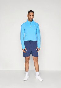 Šviesiai mėlynas ilgarankovė palaidinė su ketvirčio užtrauktuku, derinama su tamsiai mėlynais šortais. Modelis vilki baltas kojines ir sportbačius. Lygus audinys ir sportiškas kirpimas.