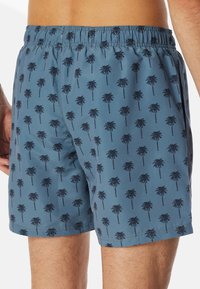 Simshorts i blått tyg med palmmönster, som har resårmidja och en avslappnad passform. Slät textur och längd till mitten av låret.