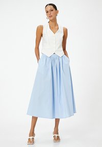 Chaleco de lino blanco con botones, acompañado de una falda recogida de color azul claro con bolsillos. La modelo lleva sandalias blancas de diseño minimalista.