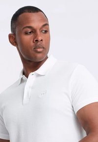 Camiseta polo blanca de tejido texturizado con una tapeta de tres botones y logo bordado en el lado izquierdo del pecho. Mangas cortas y cuello clásico.