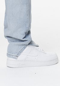 Baltās Nike Air Force 1 kedas ar perforētu purngalu, plakanām šņorēm un tekstūrētu zoli, valkātas ar gaiši ziliem džinsiem.