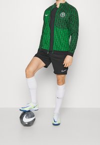Chaqueta verde con cremallera y patrones geométricos negros, pantalones cortos negros, calcetines blancos y zapatillas Nike; el balón presenta un diseño en blanco y negro.