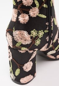 Botte noire à talon haut avec broderie florale rose et verte et une fermeture éclair noire visible sur le côté.