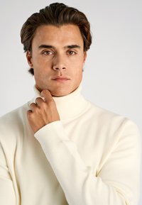 Cremefarbener Rollkragenpullover aus weichem, strukturiertem Strick. Lange Ärmel, figurbetonte Silhouette, getragen von einer Person mit dunklen Haaren, Hand am Hals.