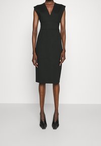 Mustapuku, jossa on V-pääntie ja hihattomat hihat, vartalonmyötäinen vyötärö ja polven mittainen helma. Valmistettu teksturoitusta kankaasta, yhdistetty mustiin korkokenkiin.