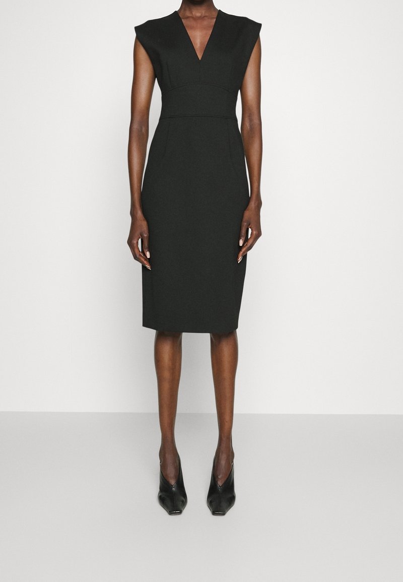 Mustapuku, jossa on V-pääntie ja hihattomat hihat, vartalonmyötäinen vyötärö ja polven mittainen helma. Valmistettu teksturoitusta kankaasta, yhdistetty mustiin korkokenkiin.