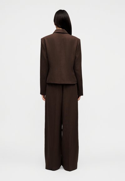 Γυναίκα με μακριά σκούρα μαλλιά, φοράει ένα καφέ σκούρο, ραμμένο σακάκι και παντελόνι με φαρδιά πόδια, στέκεται με την πλάτη προς τα εμπρός σε απλό φόντο.