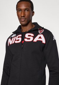 Svart zip-up-jacka med vit och röd broderad "NISSA"-logotyp och Kappa-märken. Har hög krage och slät textur.