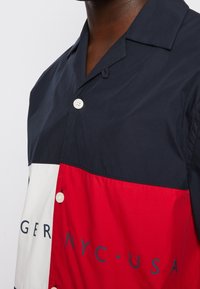 Camicia a maniche corte con bottoni, caratterizzata da blocchi di colore blu navy, rosso e bianco. Tessuto liscio con colletto classico e dettagli con logo stampato.