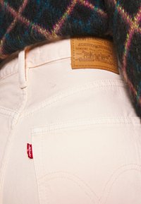Jeans de mezclilla color crema que muestran un bolsillo trasero con la etiqueta roja de Levi's y un parche de cuero marrón con el nombre y logotipo de la marca.