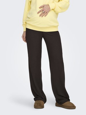 Personne portant un pantalon noir ample, des chaussons marron et un sweat-shirt jaune, avec une main posée sur l'abdomen.