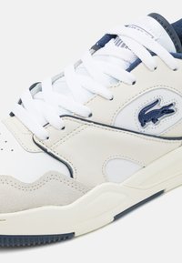 Zapatilla blanca y beige con acentos en navy, que presenta una combinación de cuero y ante, perforaciones en la puntera y un destacado logo de Lacoste.
