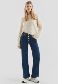 Lichtbeige fuzzy trui gecombineerd met hooggepolstelde donkerblauwe denim jeans met een rij ronde knopen; model draagt stevige witte sneakers.