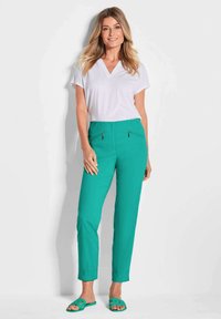 Pantalons turquoise avec poches zippées, assortis à un t-shirt blanc à col en V et des sandales vertes. Texture lisse et coupe ajustée.
