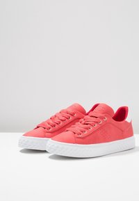Paire de baskets lacées rose corail avec semelles blanches texturées et col rembourré sur une surface blanche devant un fond gris.