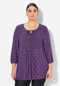 Lila Blumen-Tunika mit Rundhalsausschnitt, Schlüsselloch-Detail, 3/4-Ärmeln und elastischem Smocking, kombiniert mit dunklen Jeans.