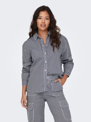 Femme aux longs cheveux foncés portant une chemise boutonnée à rayures verticales noires et blanches assortie avec un pantalon, une main dans la poche.
