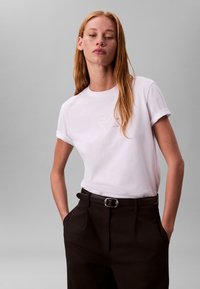 Tricou alb din bumbac cu mâneci suflate, cu un logo subtil în auriu deschis. Purtat cu pantaloni negri cu talie înaltă și o curea neagră.