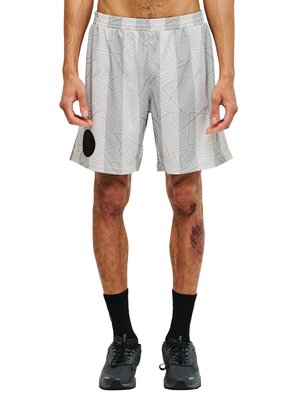 Mand iført lysegrå mønstrede sportsshorts, sorte strømper og sorte sneakers, stående med synlige skrammer på benene.