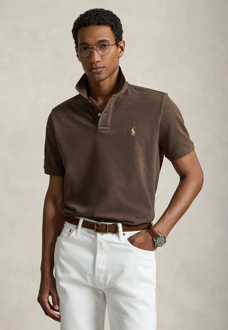 Noor mees ümarate prillidega, pruun polo särgiga, mille krae on üles keeratud, valged püksid, pruun vöö ja käekell, seisab ühe käega taskus.