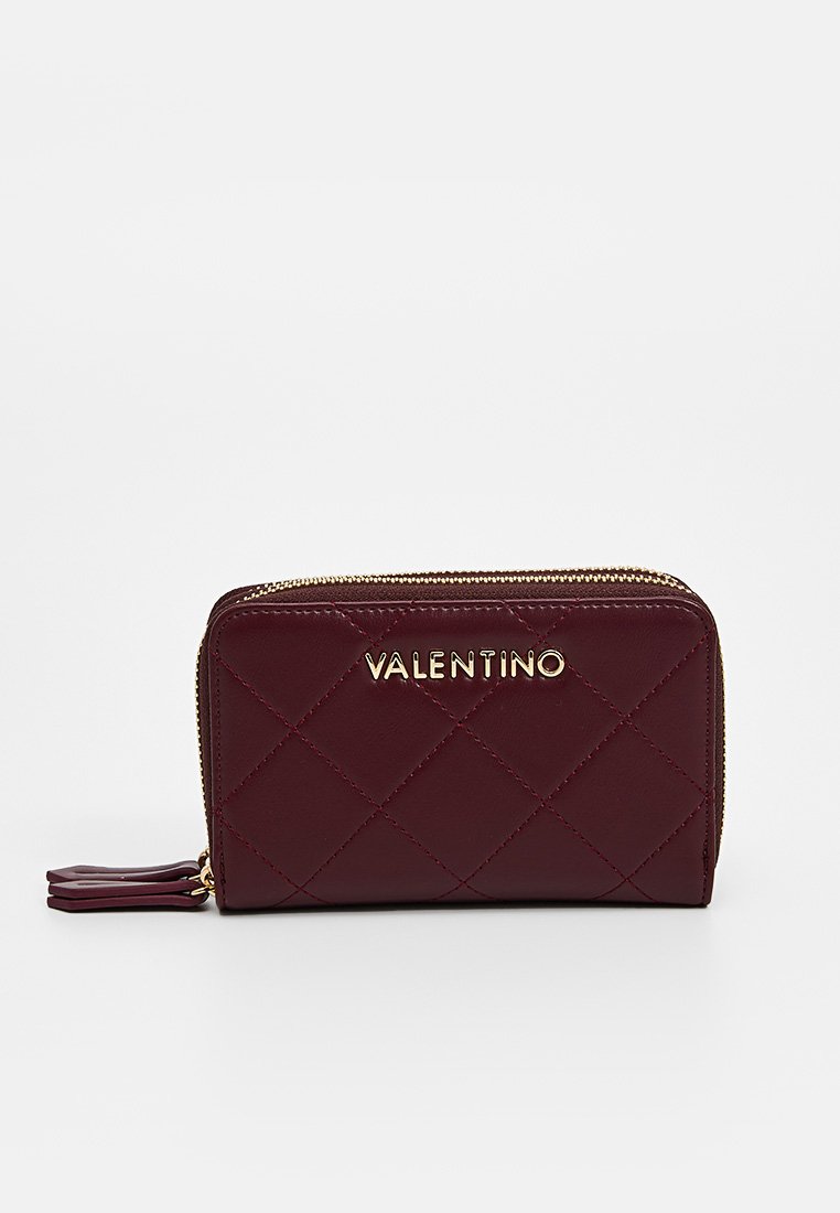Burgundowa skórzana portmonetka z pikowanym wzorem, złotym logo i zamknięciem na zamek. Posiada dekoracyjny chwost i kompaktowy prostokątny kształt.