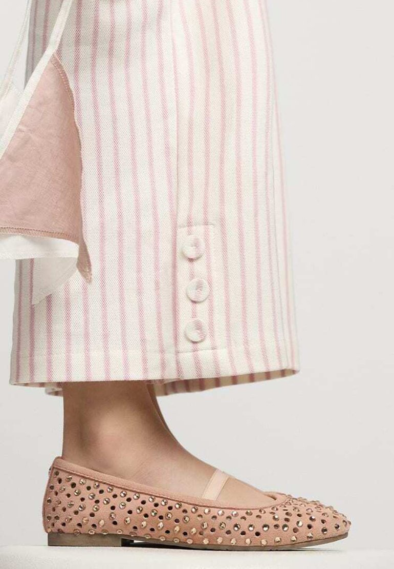 Bailarinas de charol rosa con tachuelas y una textura suave, combinadas con unos pantalones de pierna ancha de rayas verticales claras, que tienen tres botones en el dobladillo.