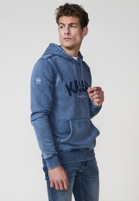 Modrá bavlnená mikina s kapucňou, prednou vreckom a kontrastným logom. Dlhé rukávy s rebrovanými manžetami.