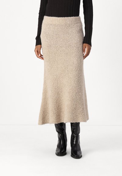 Jupe midi tricotée beige avec un motif texturé, silhouette évasée et large ceinture. Associée à des bottes noires jusqu'aux genoux.