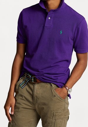 Polo morado de algodón con dos botones, con un logo verde en el pecho izquierdo. Combinado con pantalones cortos cargo color caqui y un cinturón a rayas.
