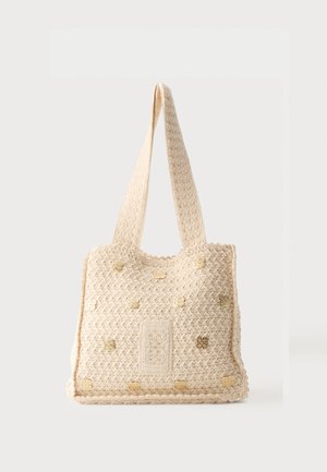 Krémefarebná tkaná taška s širokými ramennými popruhmi a malými zlatými kvetinovými dekoráciami na prednom paneli.