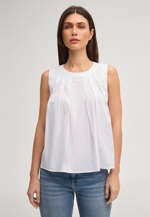 Kobieta stojąca, ubrana w białą, plisowaną bluzkę bez rękawów i niebieskie dżinsy, na neutralnym tle.