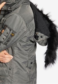 Persoon in een grijze winterjack die de mouw aanpast, met een afneembare zwarte bontvoering capuchon die aan de zijkant hangt.