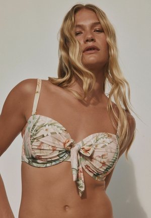 Kobieta o długich blond włosach nosi górę od bikini w kwiatowy wzór z węzłem z przodu na jednolitym jasnym tle.