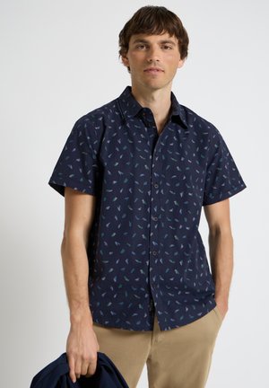 Jonge man met een marineblauw overhemd met korte mouwen en een klein blauw-groen patroon, die een marineblauw jasje in zijn linkerhand vasthoudt.