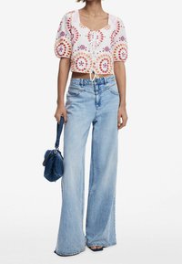 Kvinde iført en hvid cropped bluse med farverige cirkulære mønstre, lyseblå vidde jeans, holder en lille blå håndtaske.
