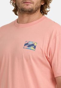 Jasnoróżowa koszulka bawełniana z okrągłym dekoltem. Posiada mały, wielokolorowy grafikę fali po lewej stronie na klatce piersiowej. Krótkie rękawy.