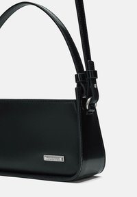 Borsa a mano in pelle nera con una forma strutturata, una tracolla removibile e una piastra logo in argento. Presenta una superficie liscia e dettagli di design minimali.